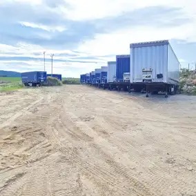 Rząd zaparkowanych naczep chłodniczych na piaszczystym placu, widoczne ślady opon, w tle zielone pola i pochmurne niebo.