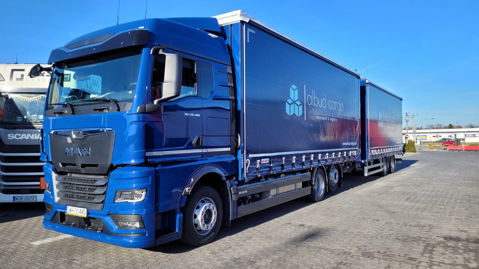 Niebieski ciągnik siodłowy MAN TGX 26.480 z naczepą, oznaczony logo 'albud cargo transport & logistic', zaparkowany na brukowanym placu, widoczny fragment innego pojazdu ciężarowego.