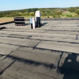 Wykończony płaski dach pokryty papą termozgrzewalną, z widocznymi rolkami papy i butlą gazową w tle, na tle zieleni.