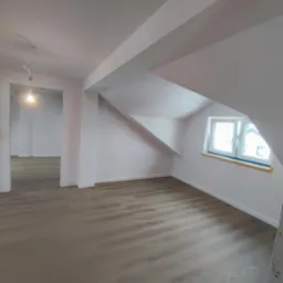 Poddasze po remoncie: świeżo pomalowane białe ściany, jasna drewniana podłoga, okno dachowe z żółtym parapetem, widoczne instalacje elektryczne.