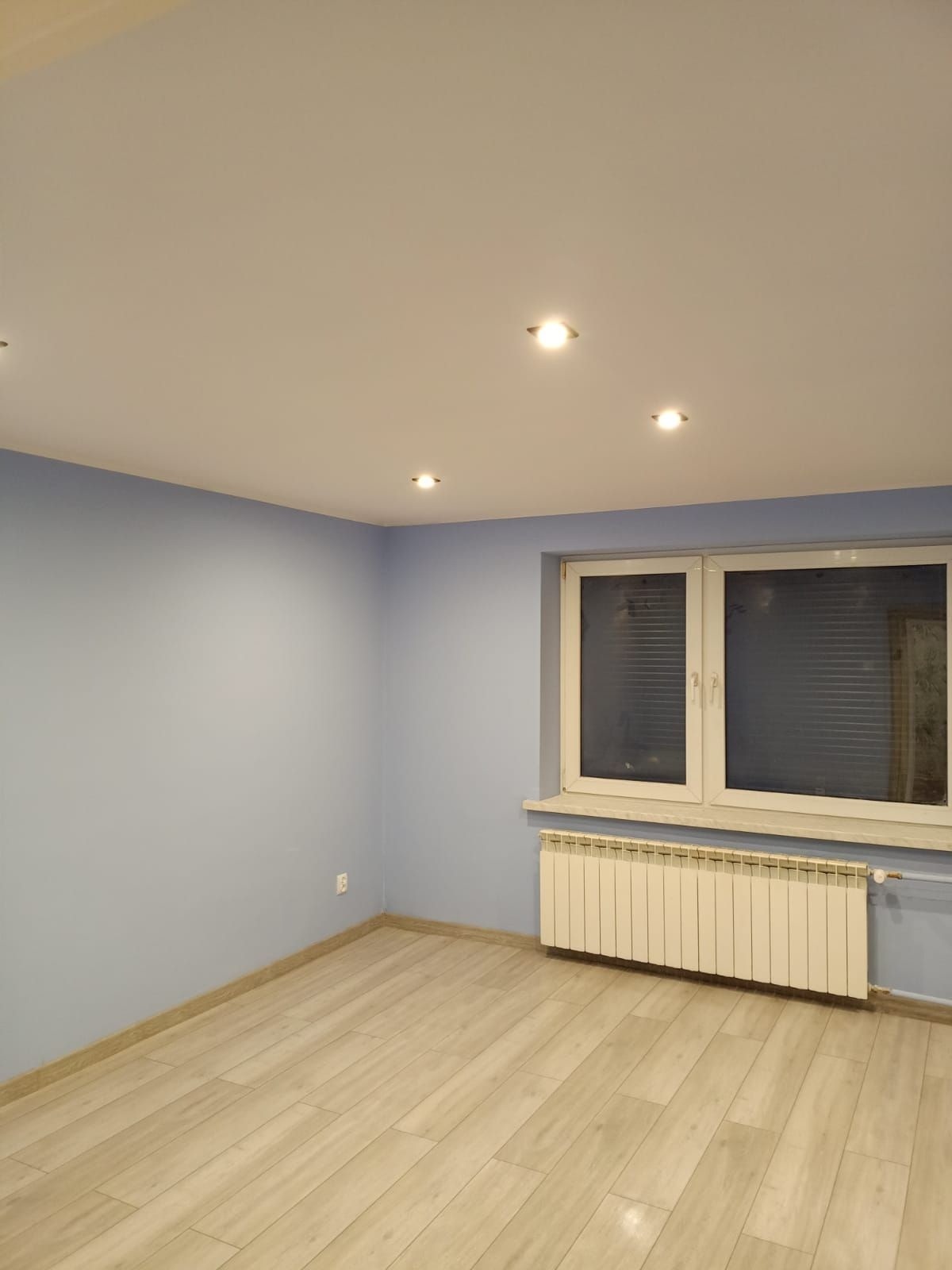 Pomalowane na jasny niebieski kolor puste pomieszczenie z drewnianą podłogą, grzejnikiem pod oknem i wbudowanymi lampami sufitowymi.
