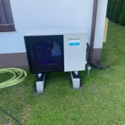 Zewnętrzna jednostka pompy ciepła marki SPRSUN ARAGO, umieszczona na betonowych bloczkach na trawniku przy ścianie budynku z brązową rynną.