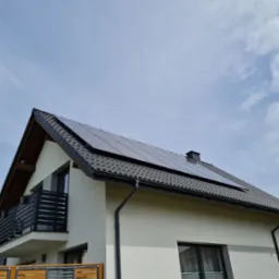 Biały dom jednorodzinny z ciemną dachówką i panelami słonecznymi na dachu, balkon z czarną balustradą, drewniane ogrodzenie w dolnej części kadru.