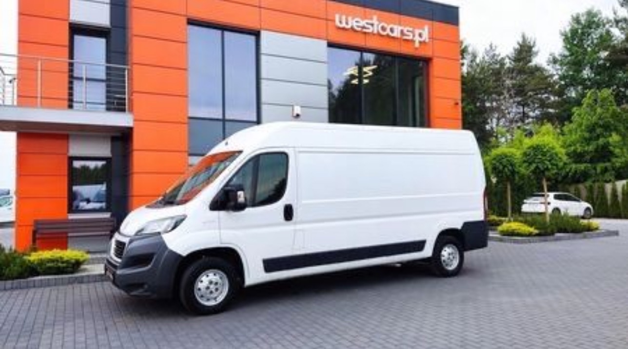 Biały furgon na tle pomarańczowo-szarego budynku z logo 'westcars.pl'. Auto zaparkowane na szarym bruku, w tle zieleń i biały samochód.