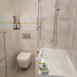 Nowoczesna łazienka z beżowymi płytkami, wiszącą toaletą, szklanym parawanem nawannowym i grzejnikiem drabinkowym. Na wannie leży opakowanie z akcesoriami montażowymi.