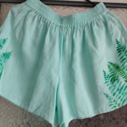 Agata Foszer - Jasnoniebieskie, bawełniane szorty z elastyczną talią i ręcznie malowanymi motywami paproci na bokach, wiszące na wieszaku na tle szklanych drzwi.