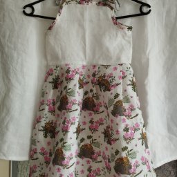 Agata Foszer - Dziecięca sukienka z białym karczkiem i wzorzystą spódnicą w kwiaty i lamparty, zawieszona na czarnym wieszaku na tle białej tkaniny.