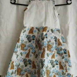 Dziecięca sukienka z białym karczkiem i wzorzystą spódnicą w panterki i niebieskie kwiaty, zawieszona na czarnym wieszaku na tle białej tkaniny.