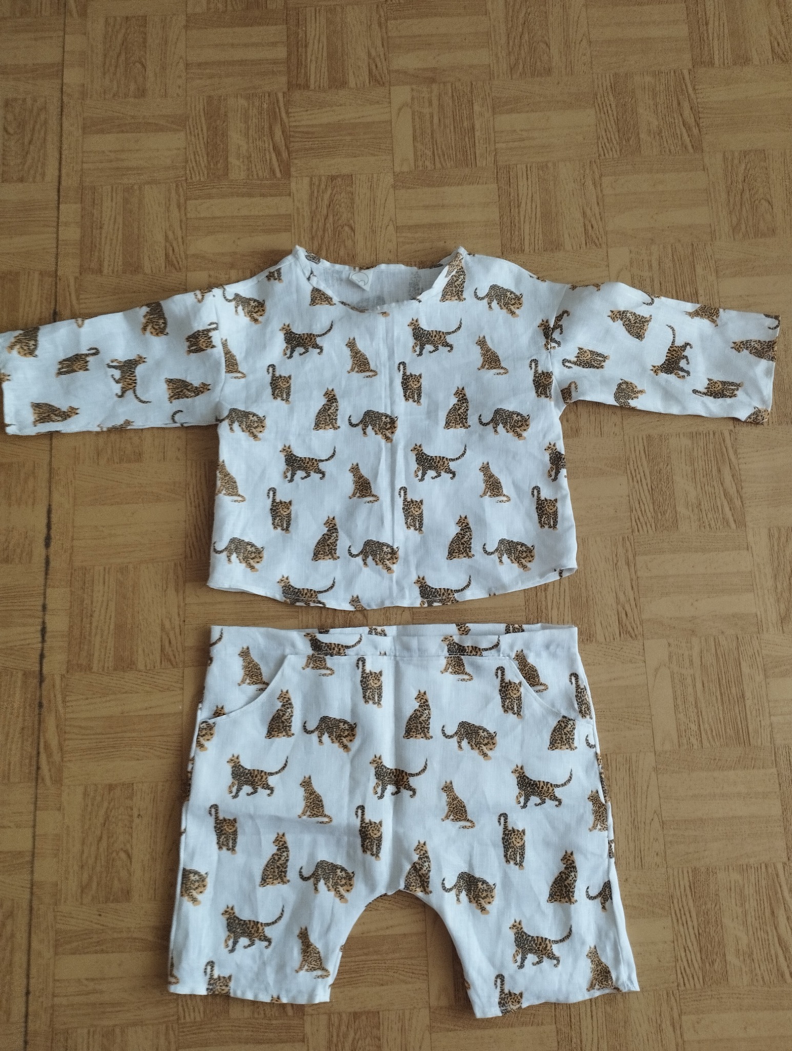 Biały komplet dziecięcy - bluzka z długim rękawem i krótkie spodenki, w brązowe koty, leżący na drewnianej podłodze.