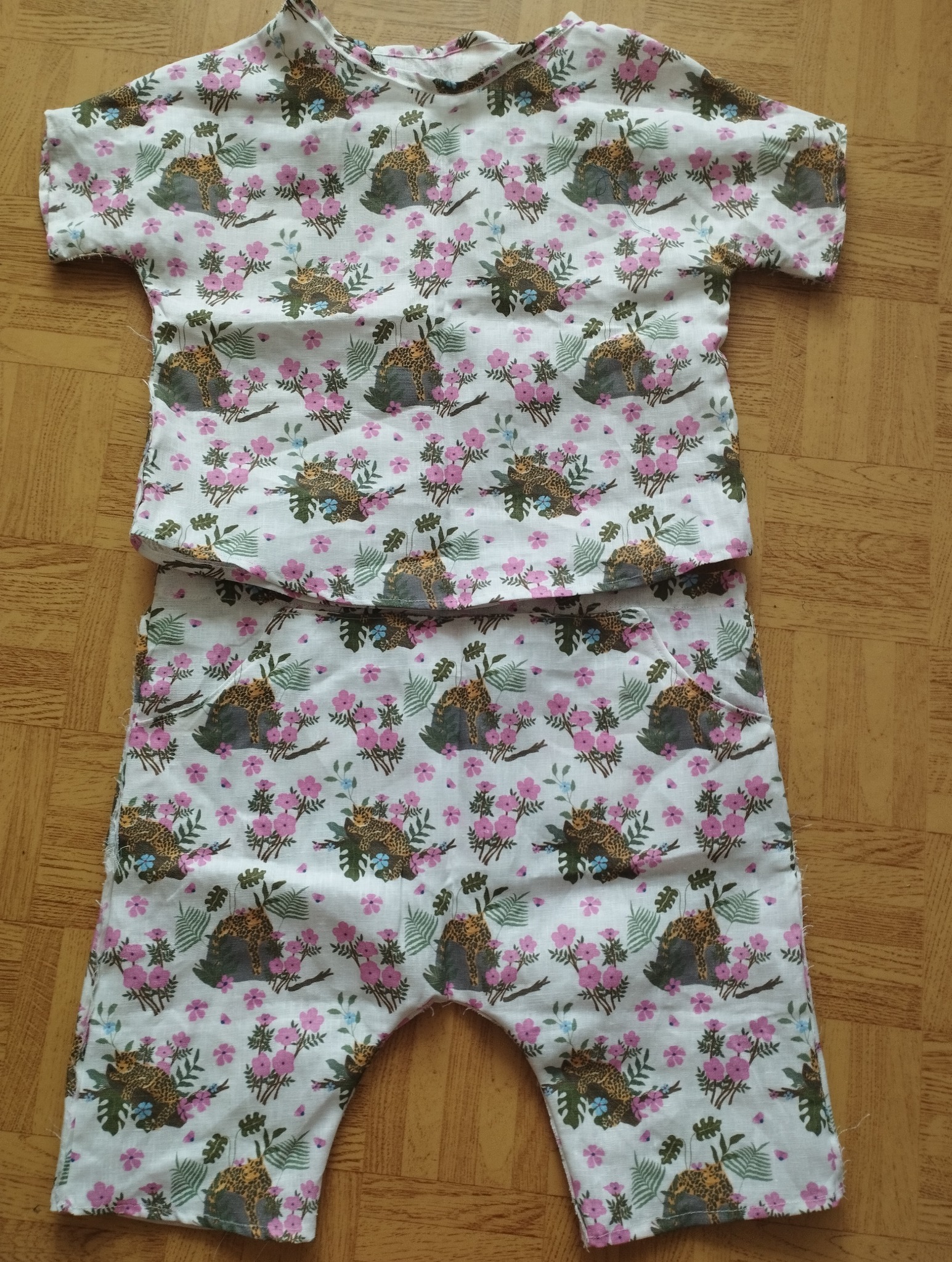 Komplet dziecięcy: krótki top i spodenki uszyte z białej tkaniny w motywy kwiatowe, liści i lampartów, leżące na drewnianej podłodze.