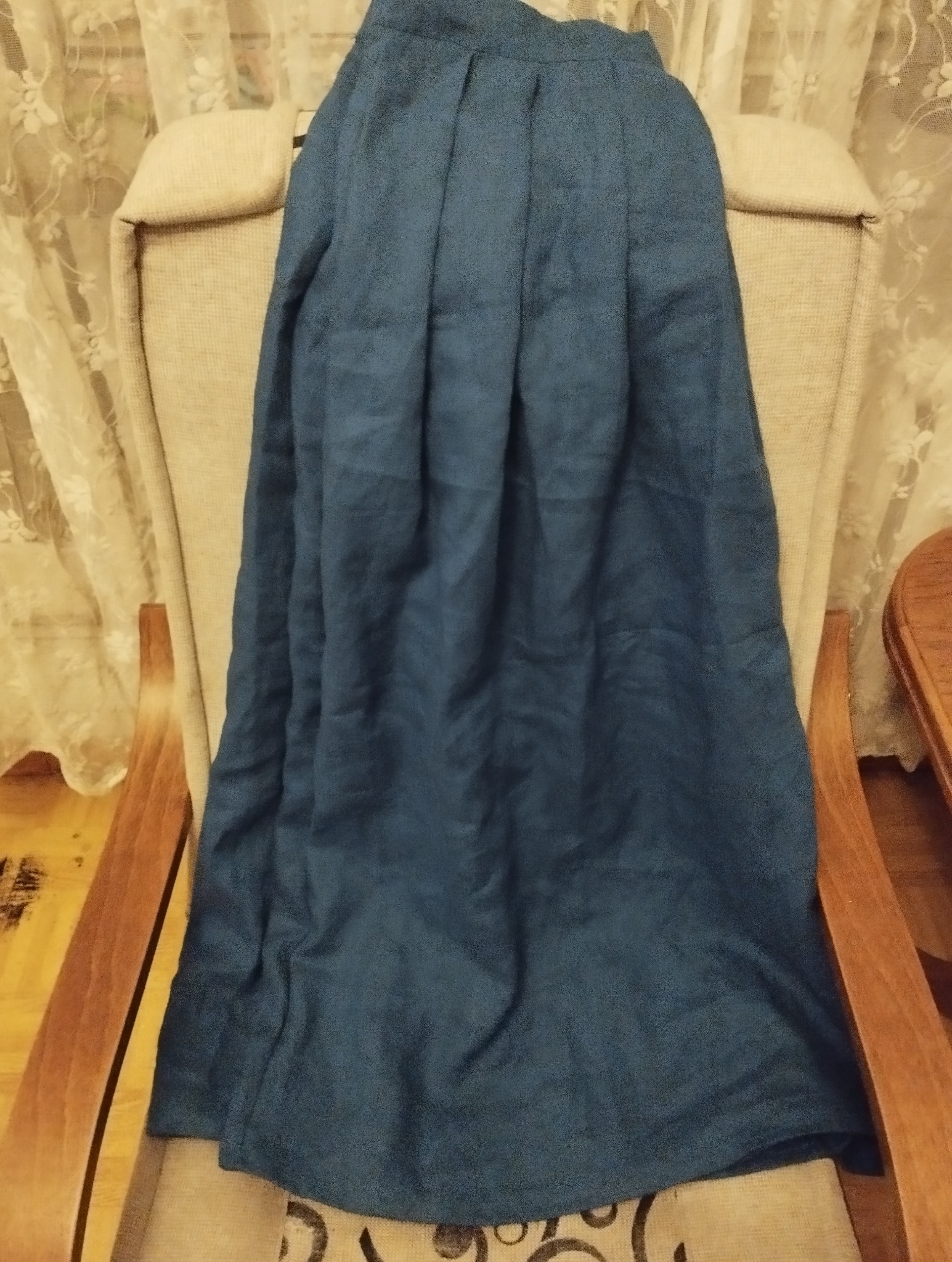 Długa, plisowana, ciemnoniebieska spódnica z lnu leży na beżowym fotelu z drewnianymi podłokietnikami, w tle firanka w kwiaty.