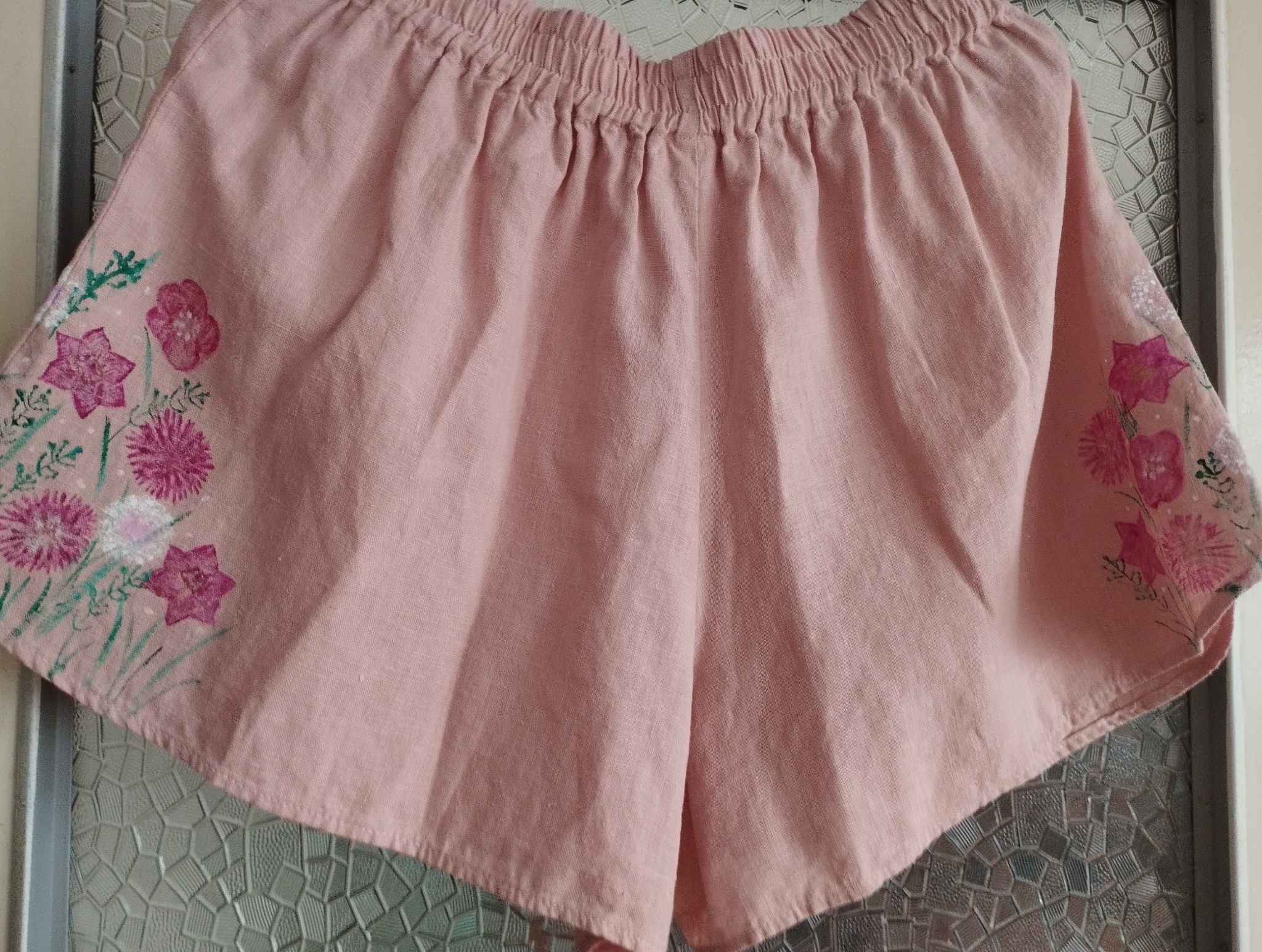 Różowe, lniane szorty z elastyczną talią, ręcznie malowane motywem kwiatowym na nogawkach, wiszące na tle wzorzystej szyby.