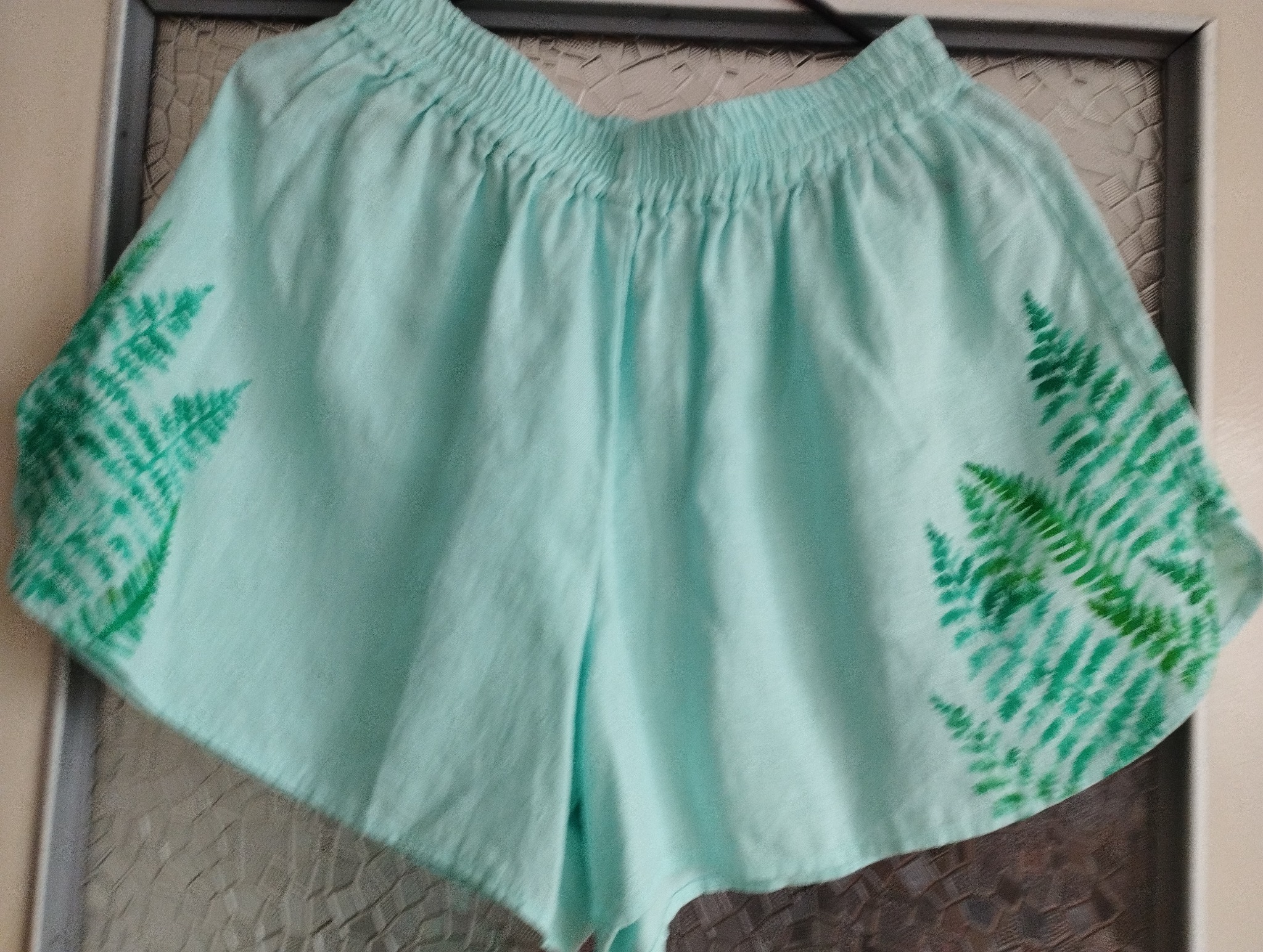 Jasnoniebieskie, bawełniane szorty z elastyczną talią i ręcznie malowanymi motywami paproci na bokach, wiszące na wieszaku na tle szklanych drzwi.