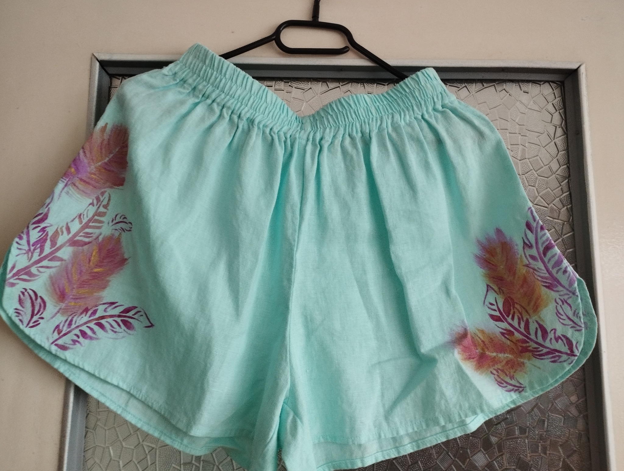 Turkusowe, lniane szorty damskie z elastyczną gumką w pasie, ozdobione ręcznie malowanymi piórami w odcieniach różu i fioletu, wiszące na czarnym wieszaku na tle wzorzystej szyby.