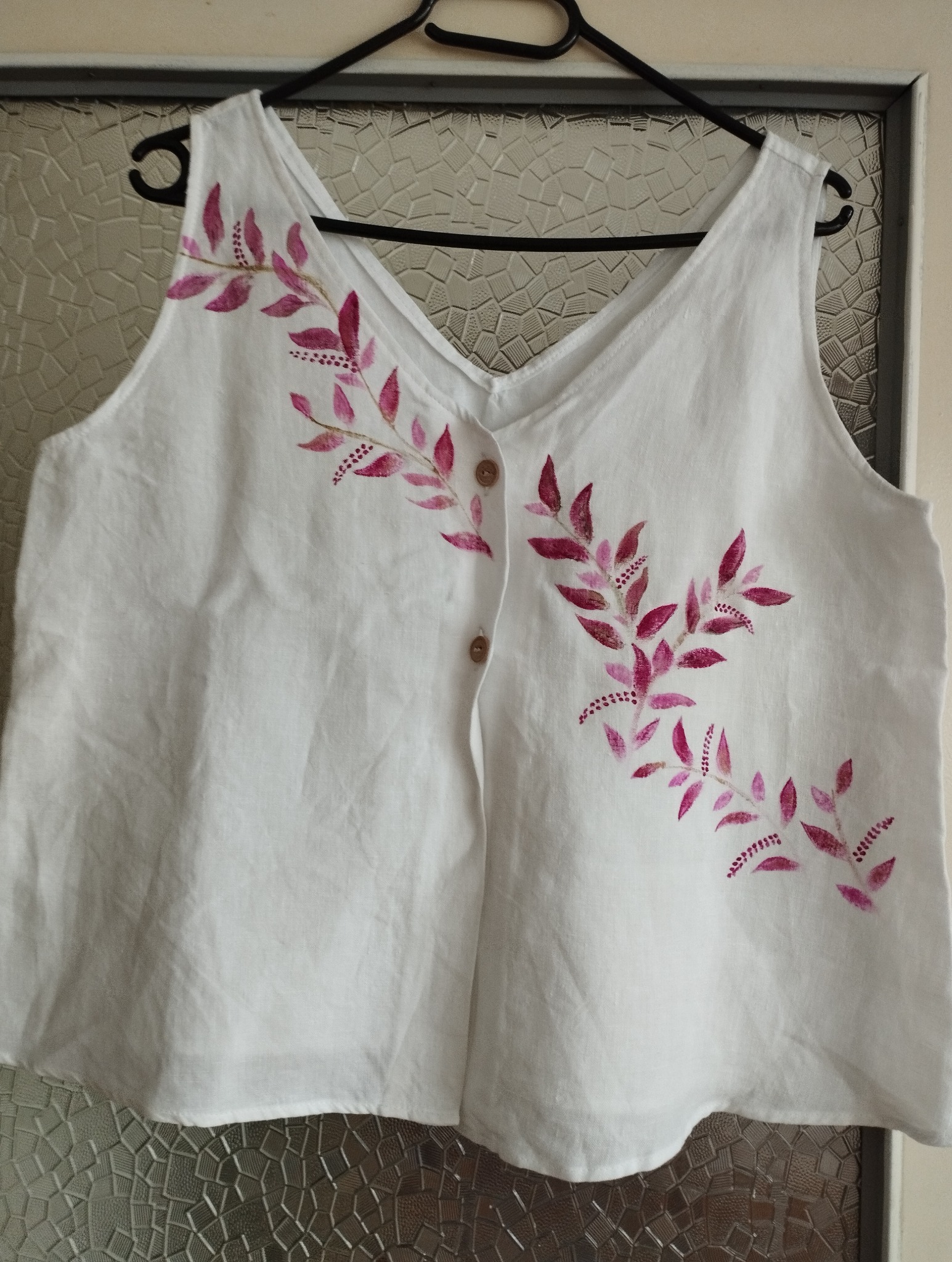 Biała, lniana bluzka na ramiączkach z ręcznie malowanymi, różowymi motywami roślinnymi, wisząca na czarnym wieszaku na tle wzorzystej szyby.