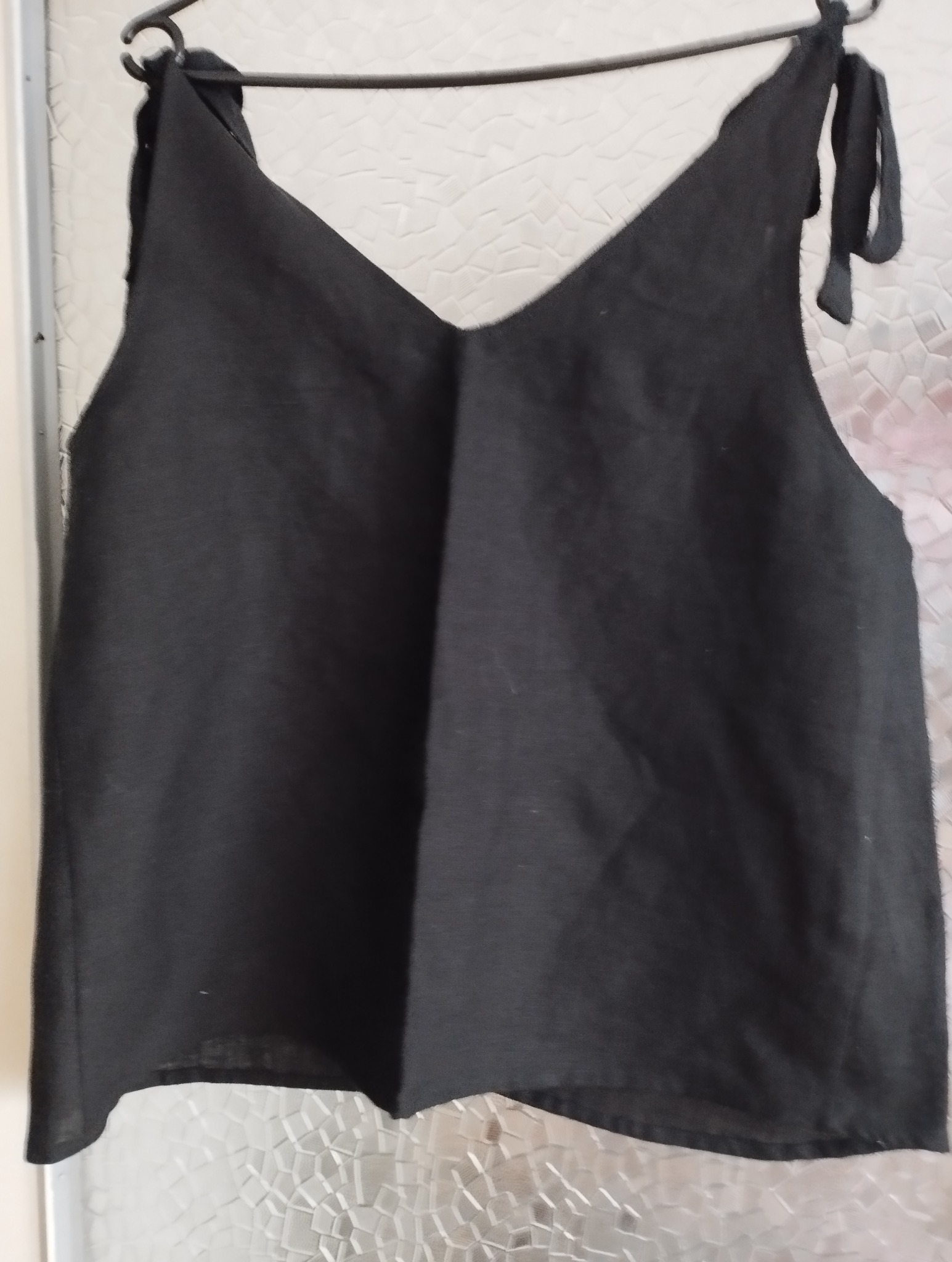 Czarna, lniana bluzka na ramiączkach z wiązaniami, wisząca na wieszaku na tle matowej szyby z wzorem.