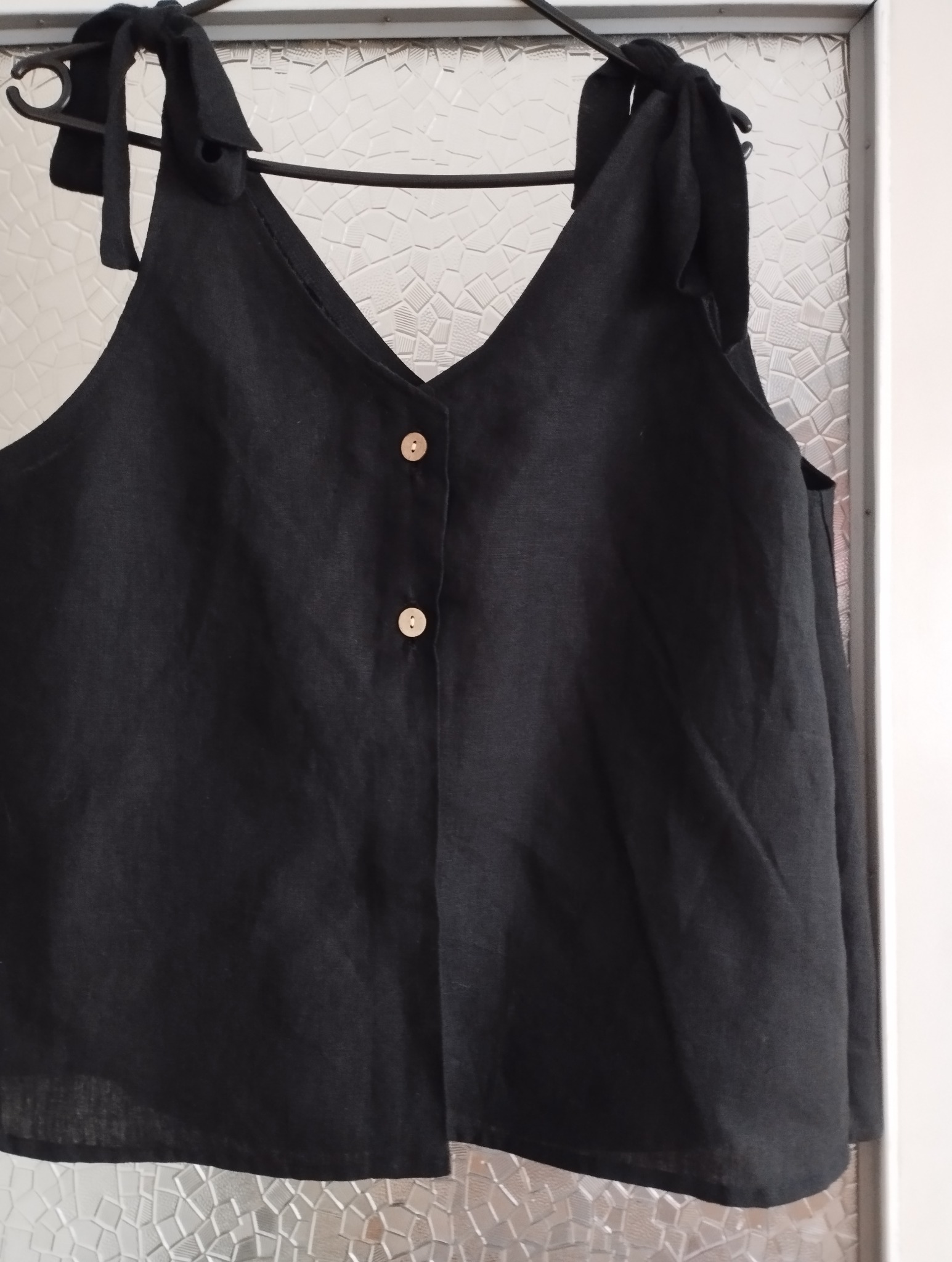 Czarna, lniana bluzka na ramiączkach z wiązaniami, zapinana na dwa drewniane guziki, wisząca na czarnym wieszaku na tle wzorzystej szyby.