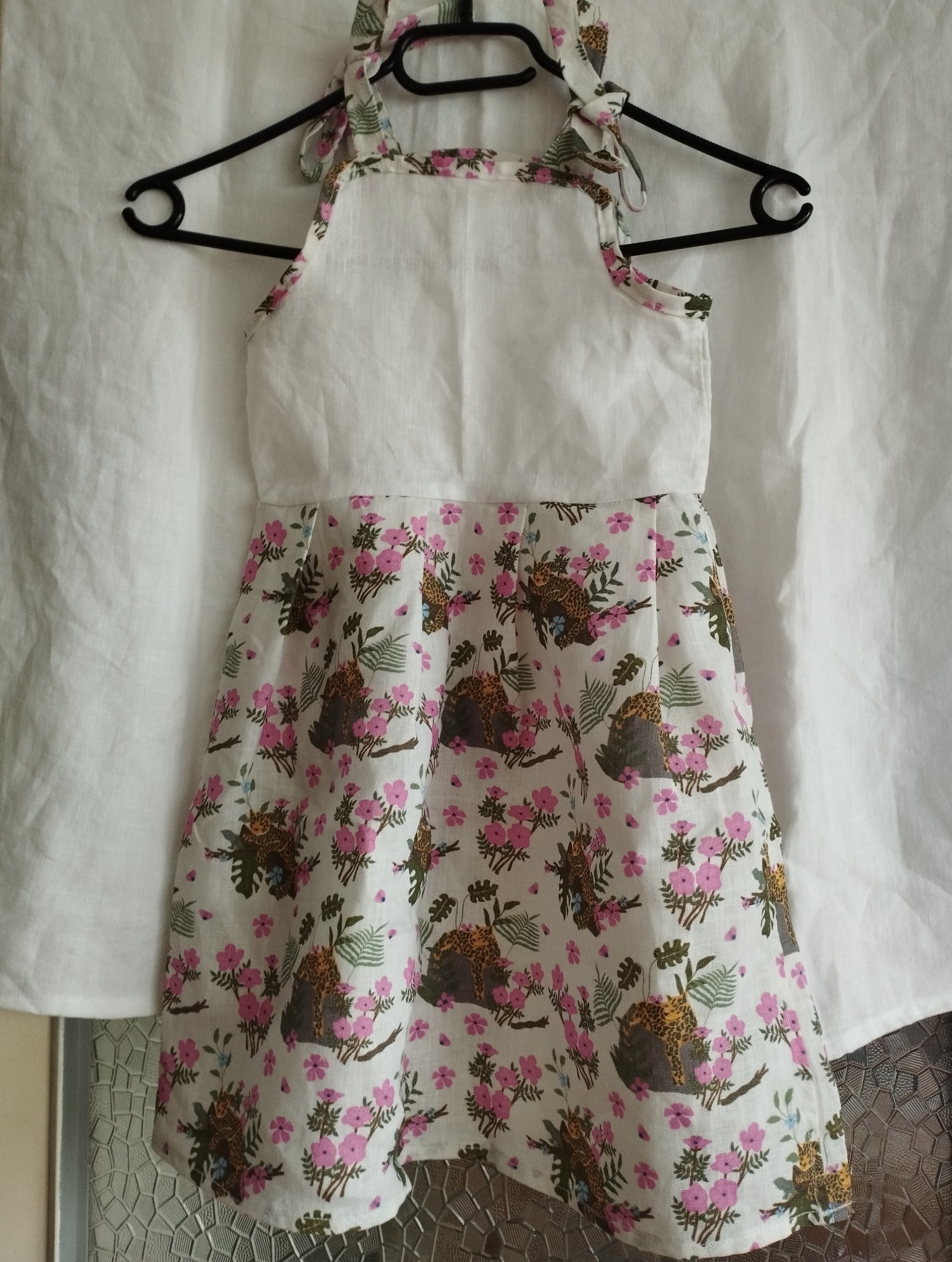 Dziecięca sukienka z białym karczkiem i wzorzystą spódnicą w kwiaty i lamparty, zawieszona na czarnym wieszaku na tle białej tkaniny.