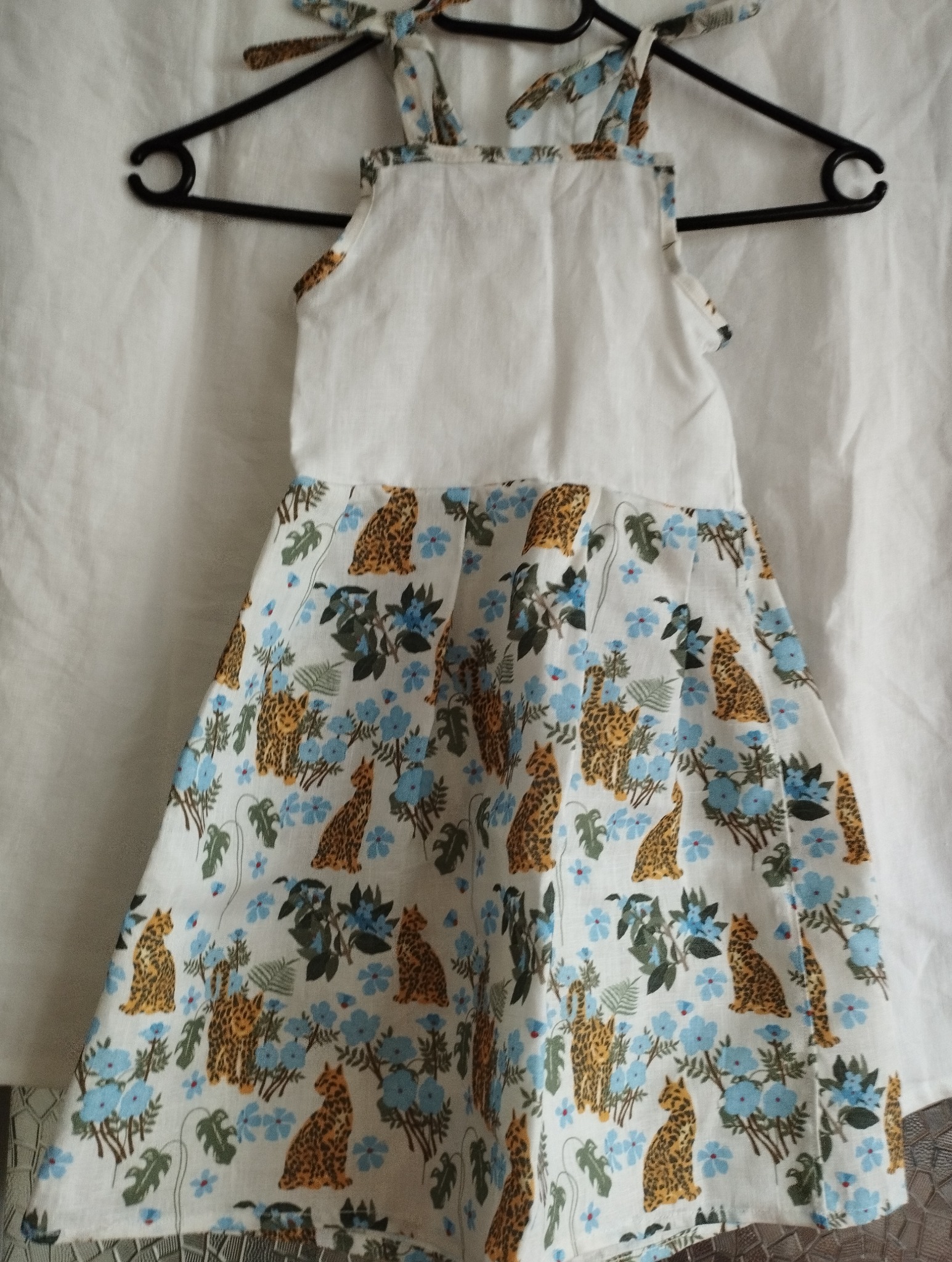 Dziecięca sukienka z białym karczkiem i wzorzystą spódnicą w panterki i niebieskie kwiaty, zawieszona na czarnym wieszaku na tle białej tkaniny.