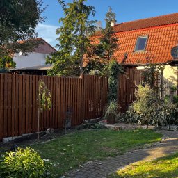 TREED - Ciemnobrązowe, pionowe deski ogrodzenia, widoczne z perspektywy ogrodu, z kwitnącą żółtą różą w pierwszym planie i domem z czerwoną dachówką w tle.