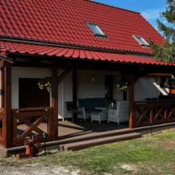 Zadaszony taras z drewnianą konstrukcją, brązowymi balustradami w kształcie krzyży, kompletem wypoczynkowym z szarej plecionki i mężczyzną w koszulce Coca-Cola pracującym w ogrodzie