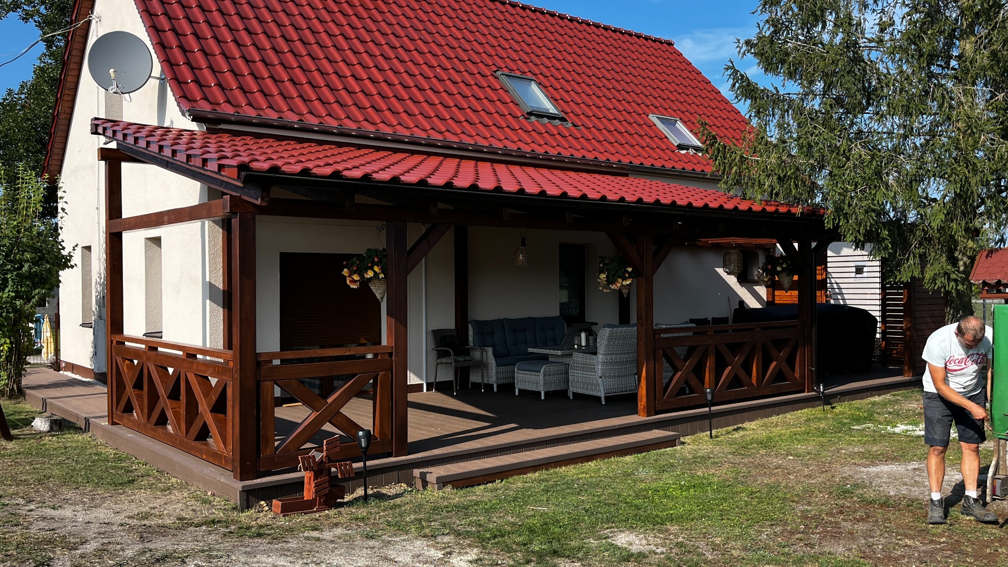 Zadaszony taras z drewnianą konstrukcją, brązowymi balustradami w kształcie krzyży, kompletem wypoczynkowym z szarej plecionki i mężczyzną w koszulce Coca-Cola pracującym w ogrodzie