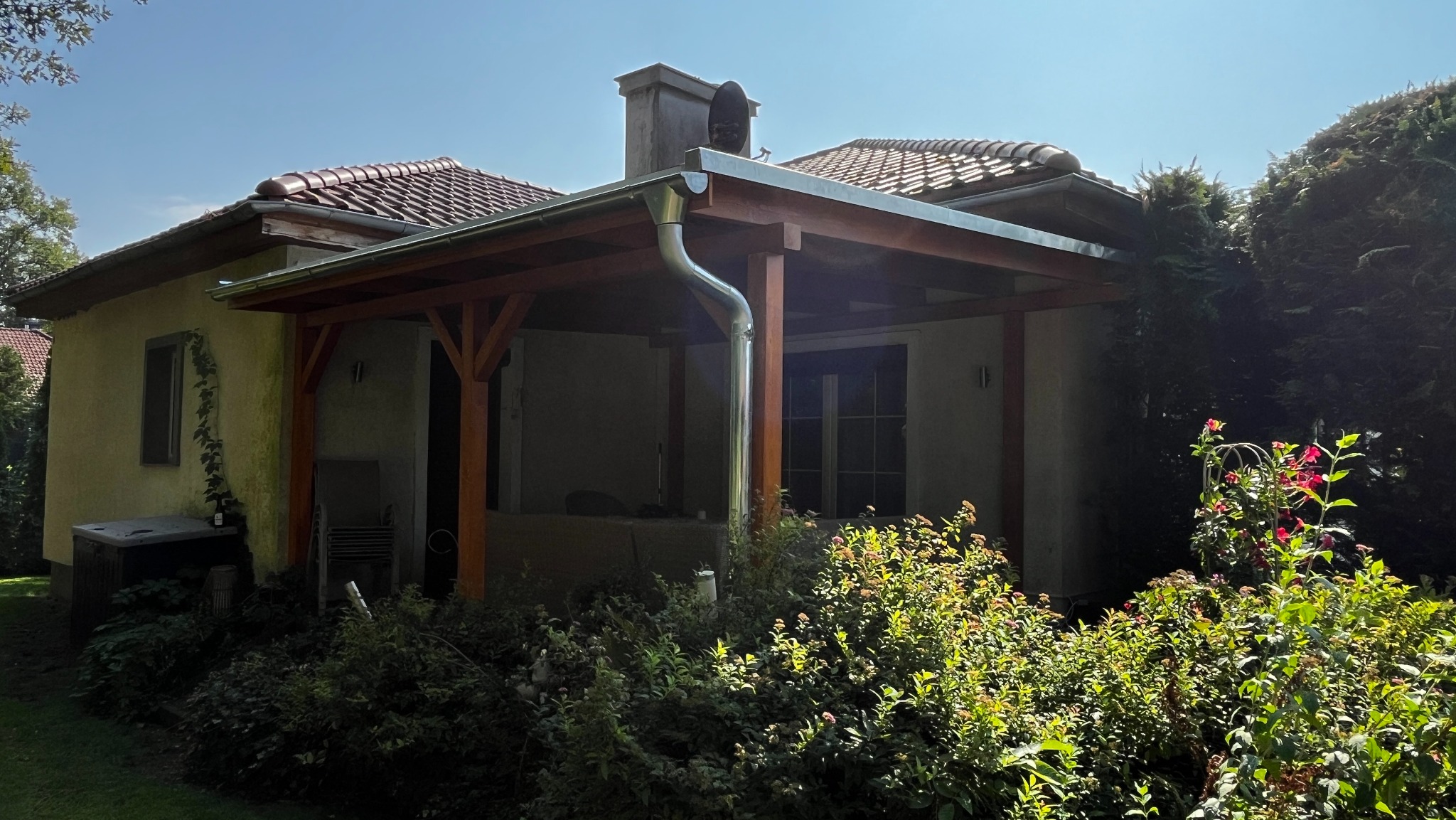 Zadaszony taras drewniany z rynną i widocznym kominem, otoczony zielenią, w słoneczny dzień.