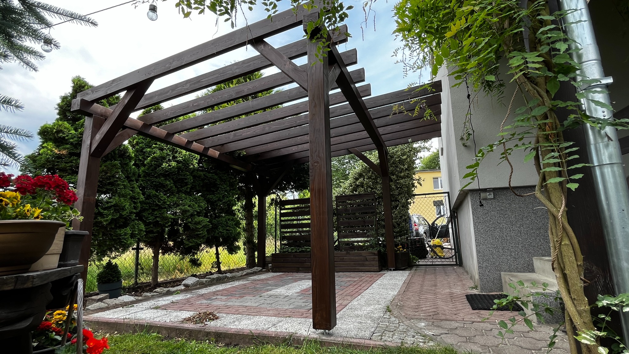 Ciemnobrązowa konstrukcja z belek tworząca zadaszenie w ogrodzie, widoczne z poziomu gruntu, z elementami dekoracyjnymi w postaci roślin pnących i donic z kwiatami, na tle domu z szarą elewacją.