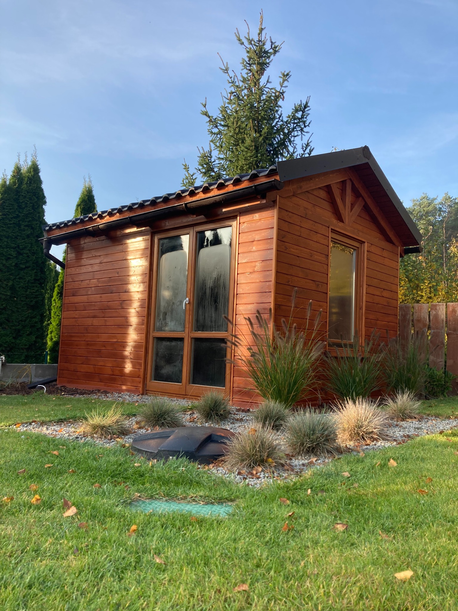 Drewniany domek narzędziowy z dużymi oknami, otoczony ozdobnymi trawami i kamieniami na trawniku, z widocznym włazem do szamba w tle.