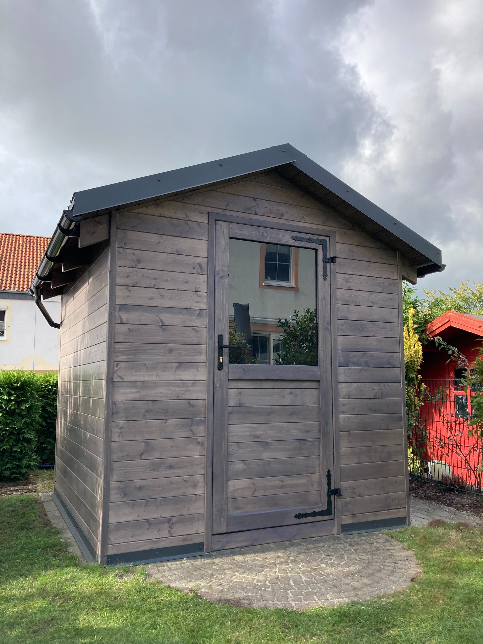 Drewniany domek narzędziowy z przeszklonymi drzwiami, pomalowany na szaro, z czarnym dachem i rynnami, stojący na trawniku z kostki brukowej, z widocznym odbiciem budynku w szybie.