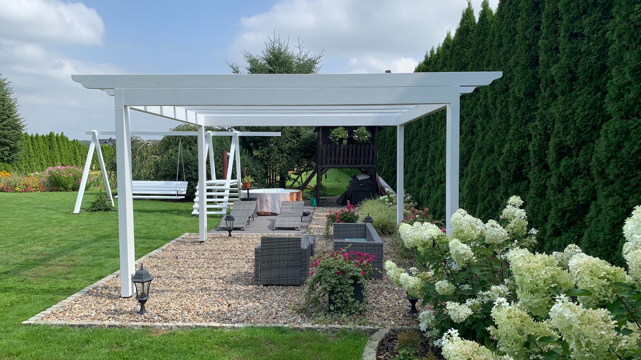 Biała pergola z huśtawką i strefą relaksu z meblami ogrodowymi na żwirowym podłożu, w tle drewniany domek dla dzieci i rząd wysokich tuj.