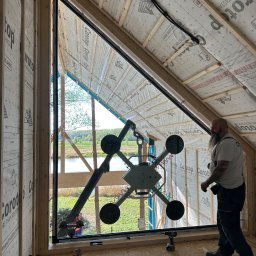 Budvar Centrum Zgierz - Montaż okna trapezowego z widokiem na jezioro. Czarny profil okienny, manipulator próżniowy w użyciu, widoczna osoba w tle i narzędzia. Budowa w stanie surowym.