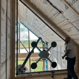 Montaż okna trapezowego z widokiem na jezioro. Czarny profil okienny, manipulator próżniowy w użyciu, widoczna osoba w tle i narzędzia. Budowa w stanie surowym.