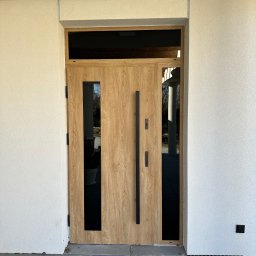 Budvar Centrum Zgierz - Nowoczesne drzwi wejściowe z drewnianym wykończeniem i czarnymi, pionowymi przeszkleniami, zamontowane w jasnej elewacji budynku. Minimalistyczny design.