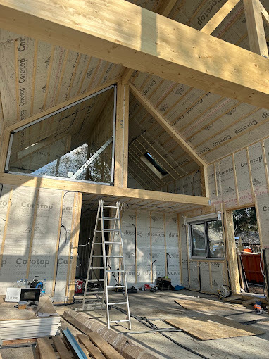 Montaż okna w surowym wnętrzu domu z drewnianą konstrukcją. Widoczna drabina i materiały budowlane. Okno górne, trójkątne, z widokiem na zewnątrz.