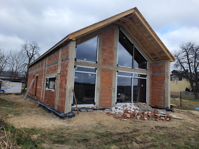 Surowy stan domu z czerwonej cegły z zamontowanymi oknami z białymi ramami; widoczne okna trójkątne na poddaszu i przesuwne okno na parterze.