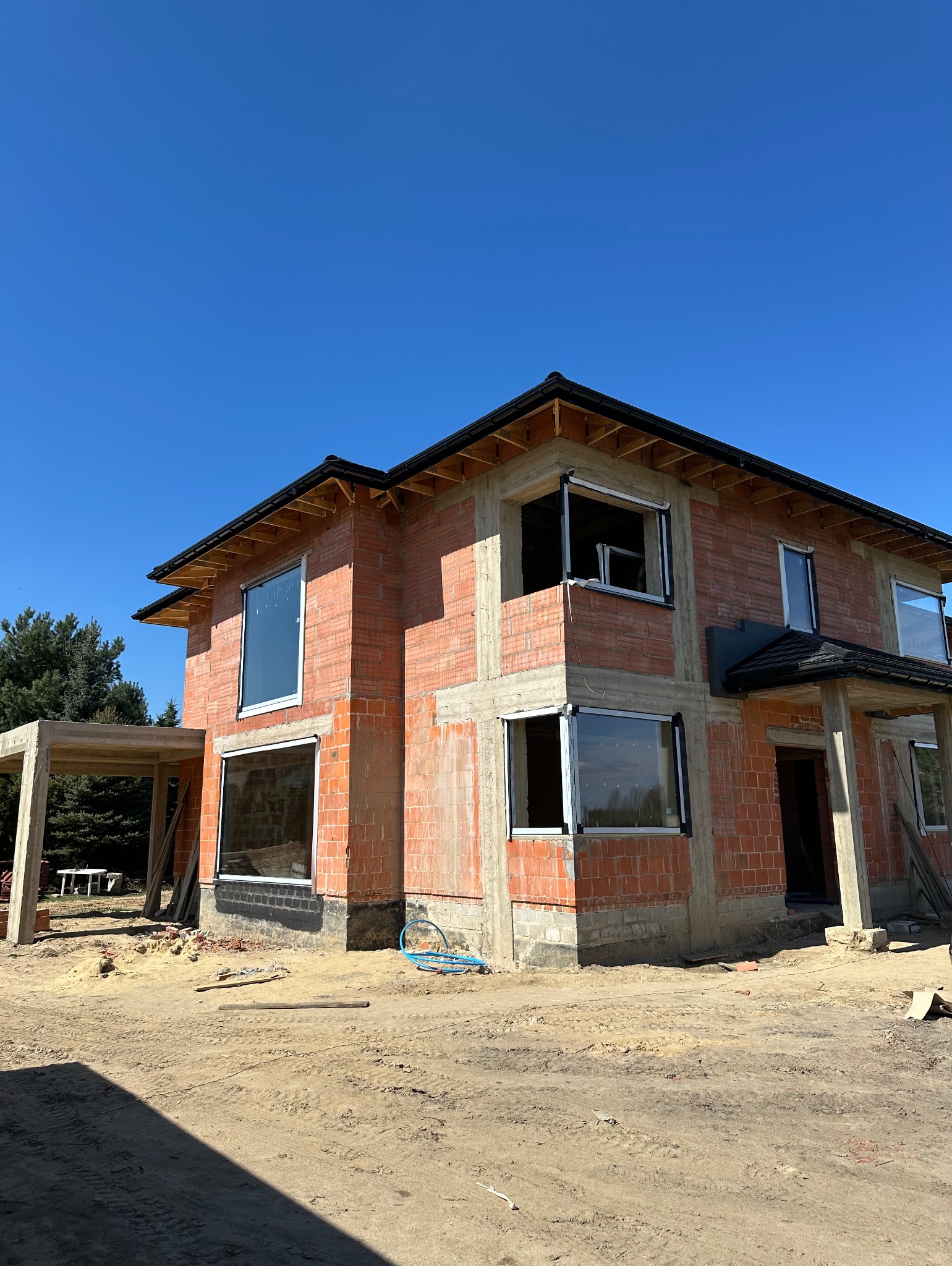 Surowy dwupiętrowy dom z czerwonej cegły w trakcie budowy, z zamontowanymi oknami w białych ramach, na tle błękitnego nieba.