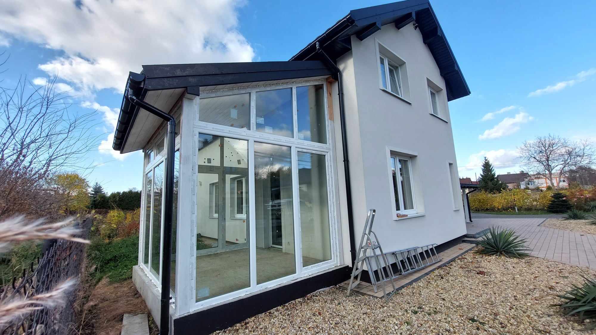 Biała, przeszklona weranda z widocznymi ramami okiennymi, dobudowana do domu. Drabina oparta o ścianę. Błękitne niebo z chmurami odbija się w szybach.