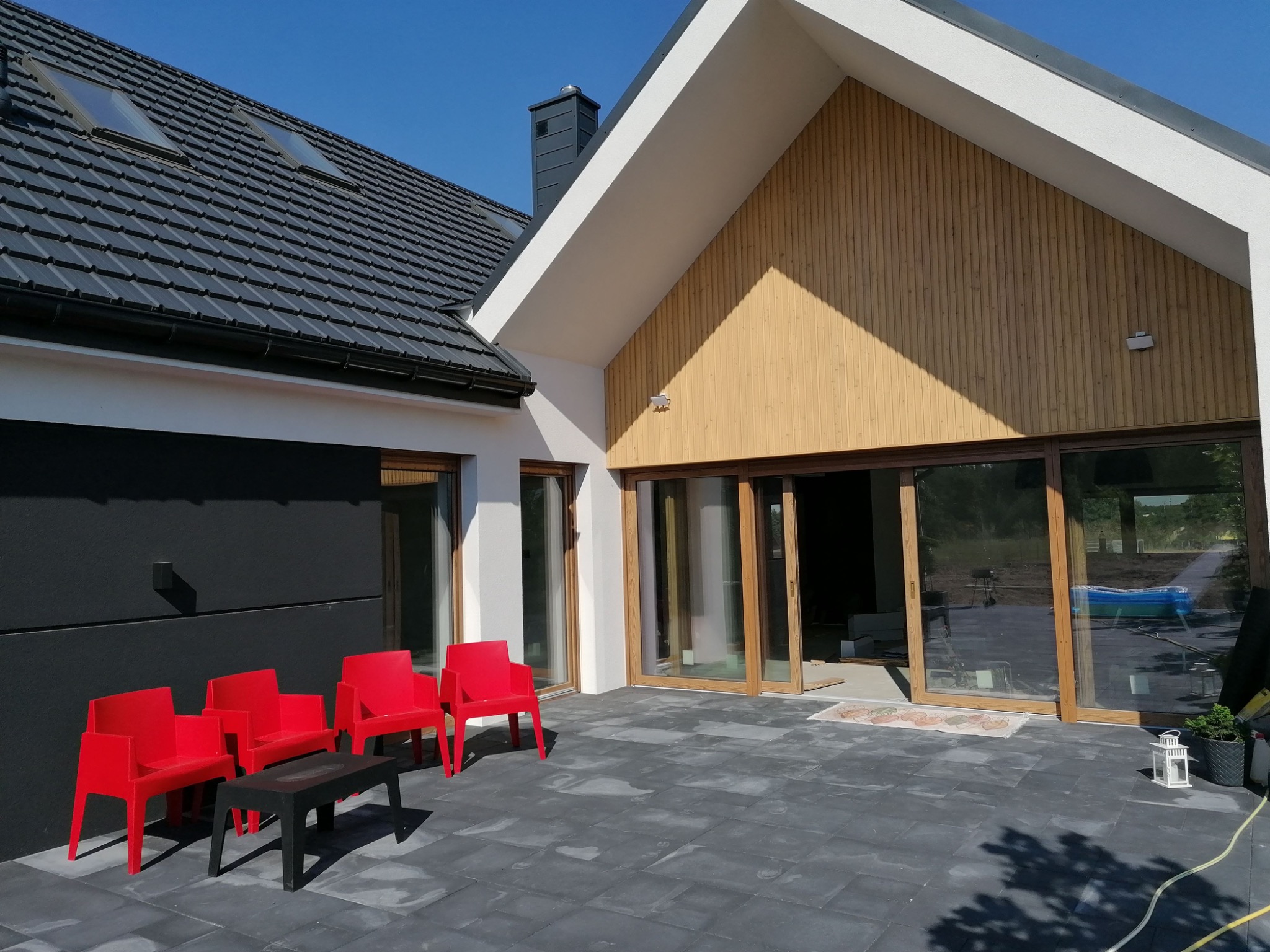 Nowoczesny dom z czarnym dachem i jasną elewacją. Widoczne duże przesuwne okna tarasowe, cztery czerwone krzesła i stolik na szarym tarasie.