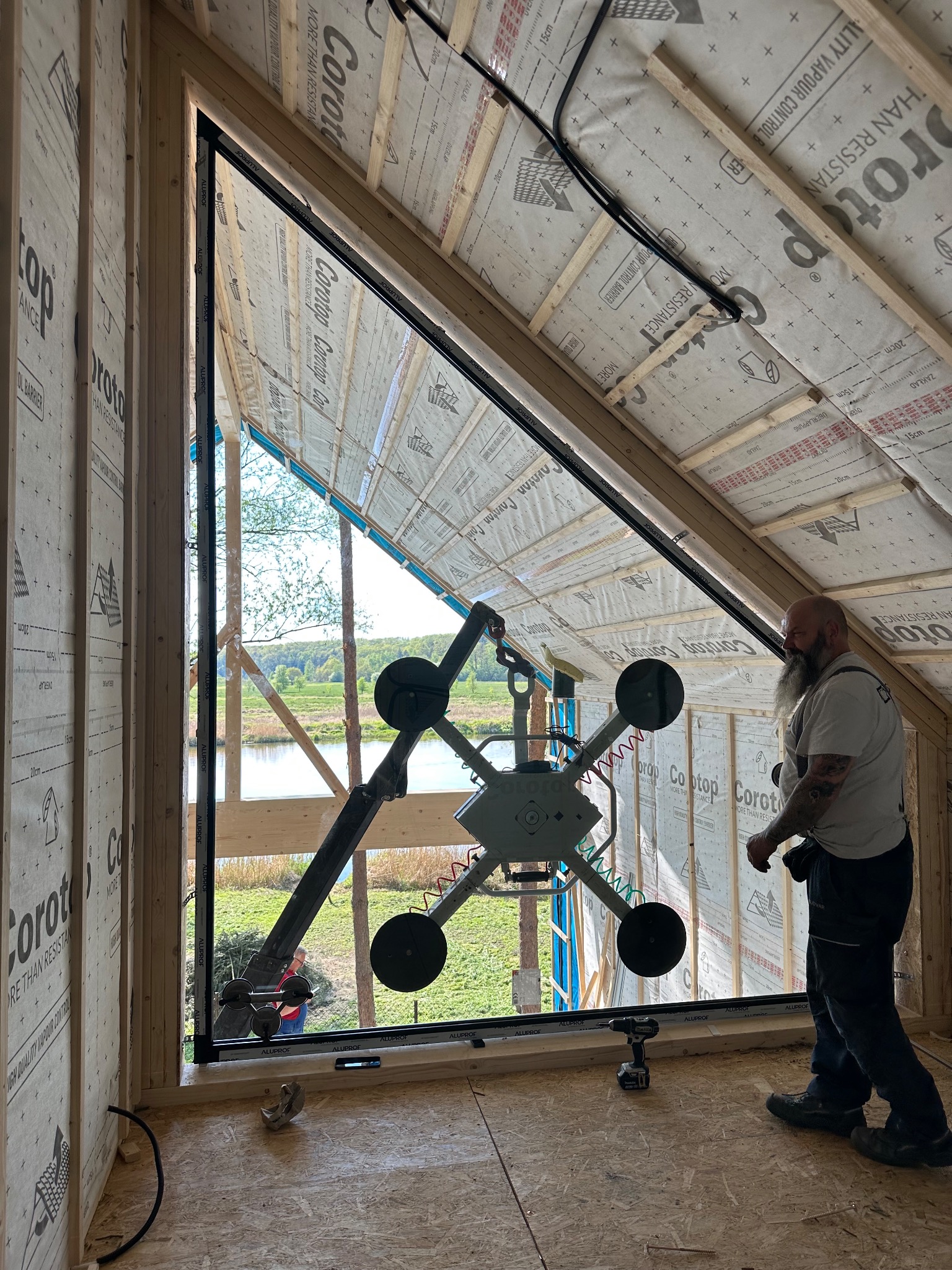 Montaż okna trapezowego z widokiem na jezioro. Czarny profil okienny, manipulator próżniowy w użyciu, widoczna osoba w tle i narzędzia. Budowa w stanie surowym.