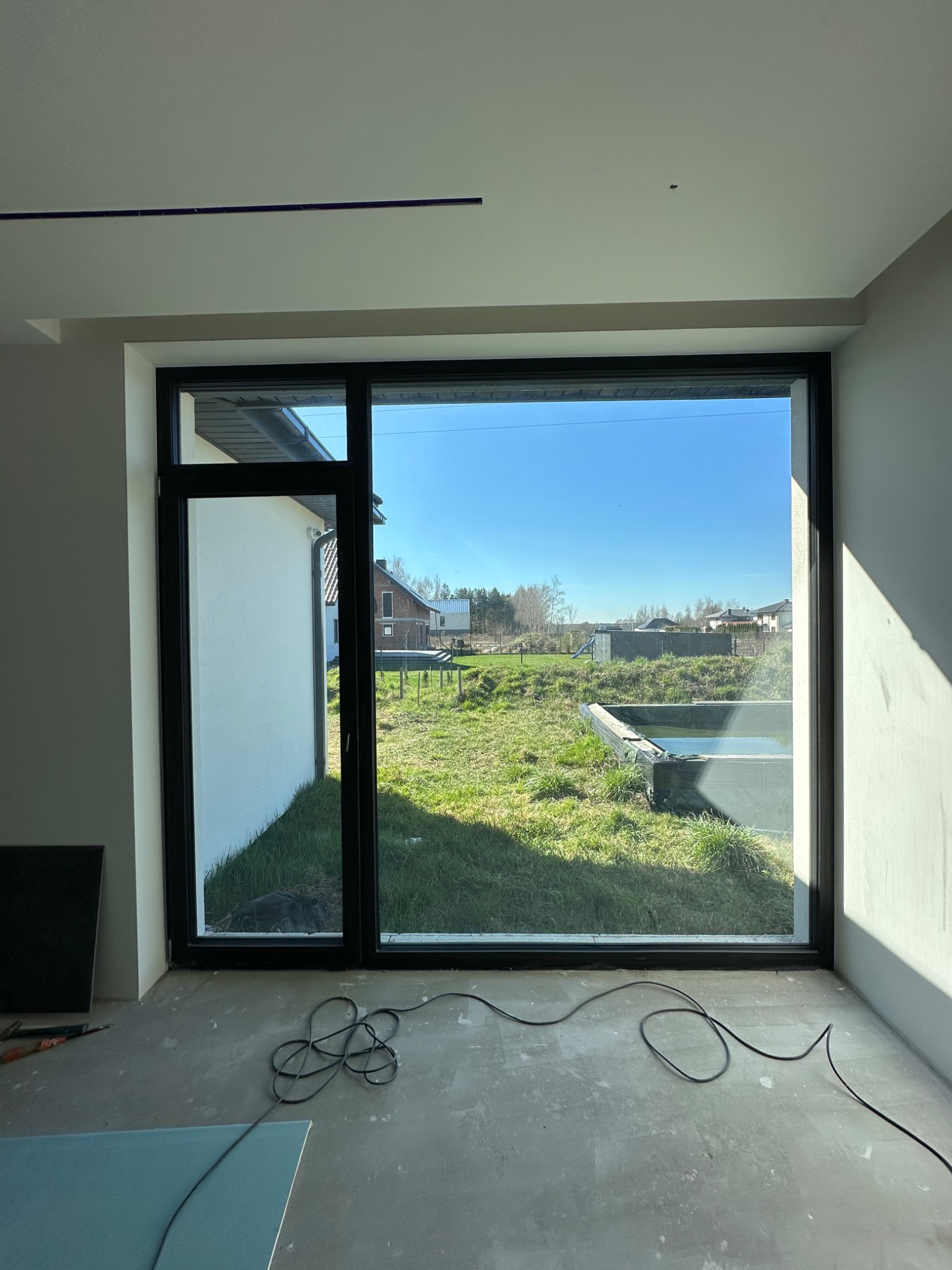 Duże okno z czarną ramą w surowym wnętrzu, widok na zewnątrz na trawnik i domy. Kabel na podłodze, panele obok okna. Dobre doświetlenie wnętrza.