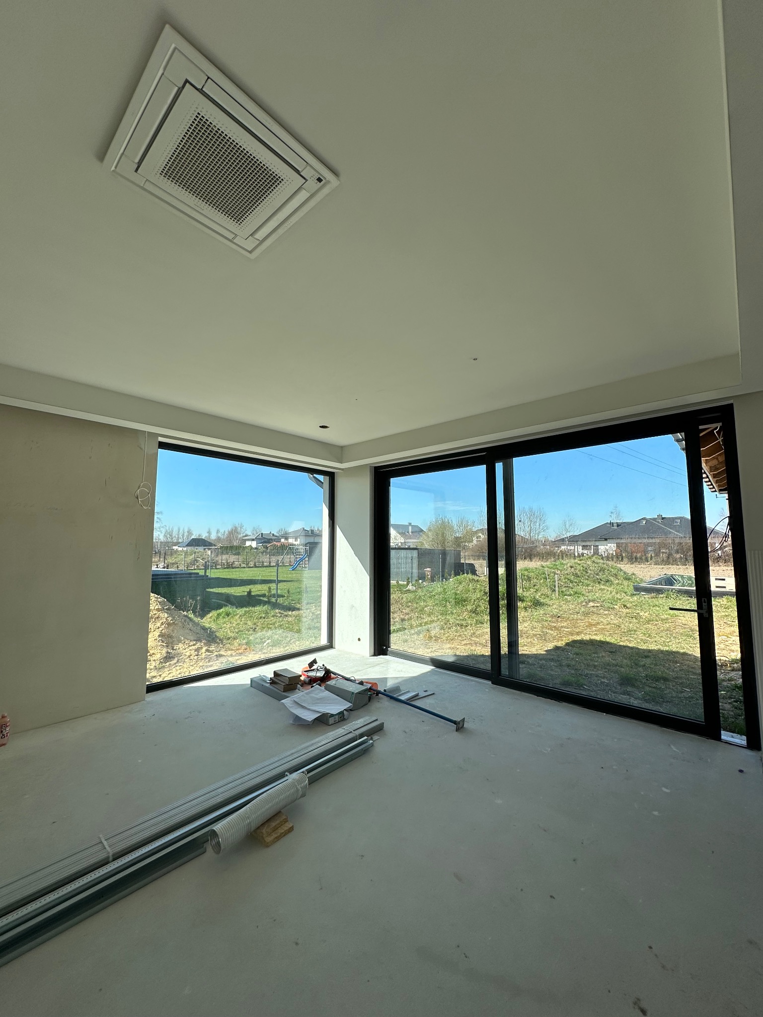Wnętrze domu w trakcie wykańczania z dużymi oknami aluminiowymi przesuwnymi z widokiem na zewnątrz. Na podłodze leżą narzędzia i materiały budowlane.