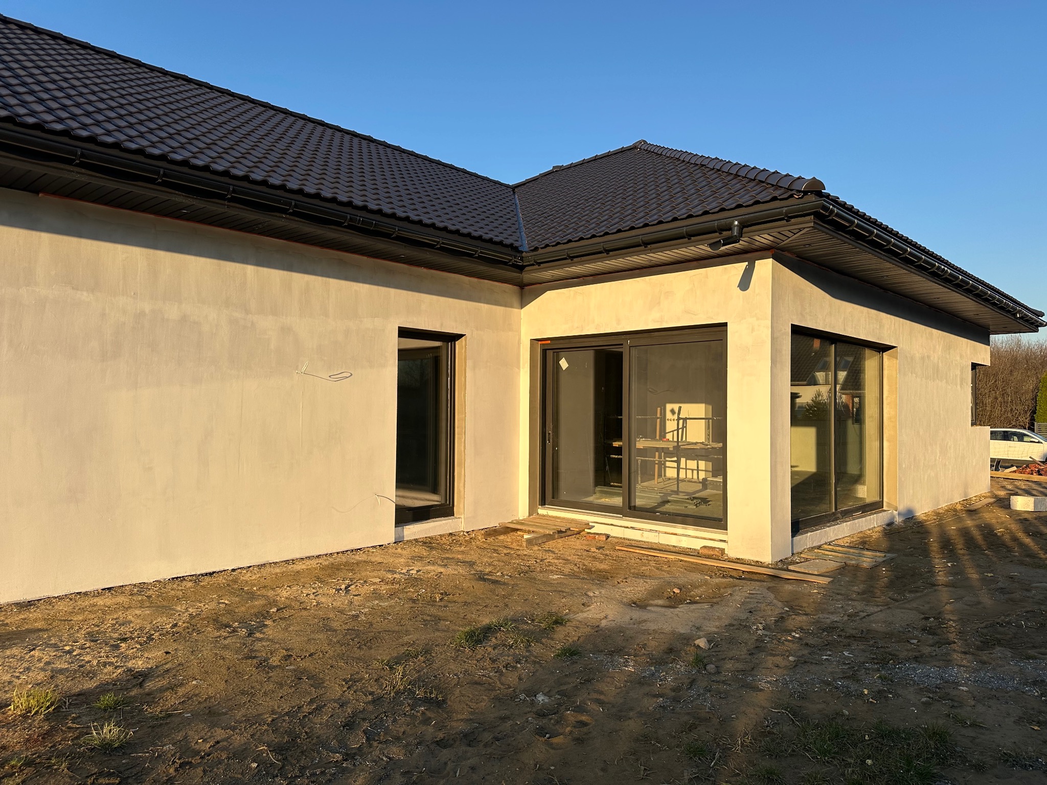 Dom jednorodzinny z grafitową dachówką i dużymi oknami przesuwnymi, w trakcie wykończenia elewacji zewnętrznej. Wnętrze widoczne przez okna.