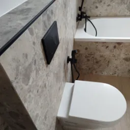 Wykończona łazienka z beżowymi płytkami imitującymi marmur, wisząca toaleta z czarnym przyciskiem spłukującym i czarna bateria bidetowa obok wanny.