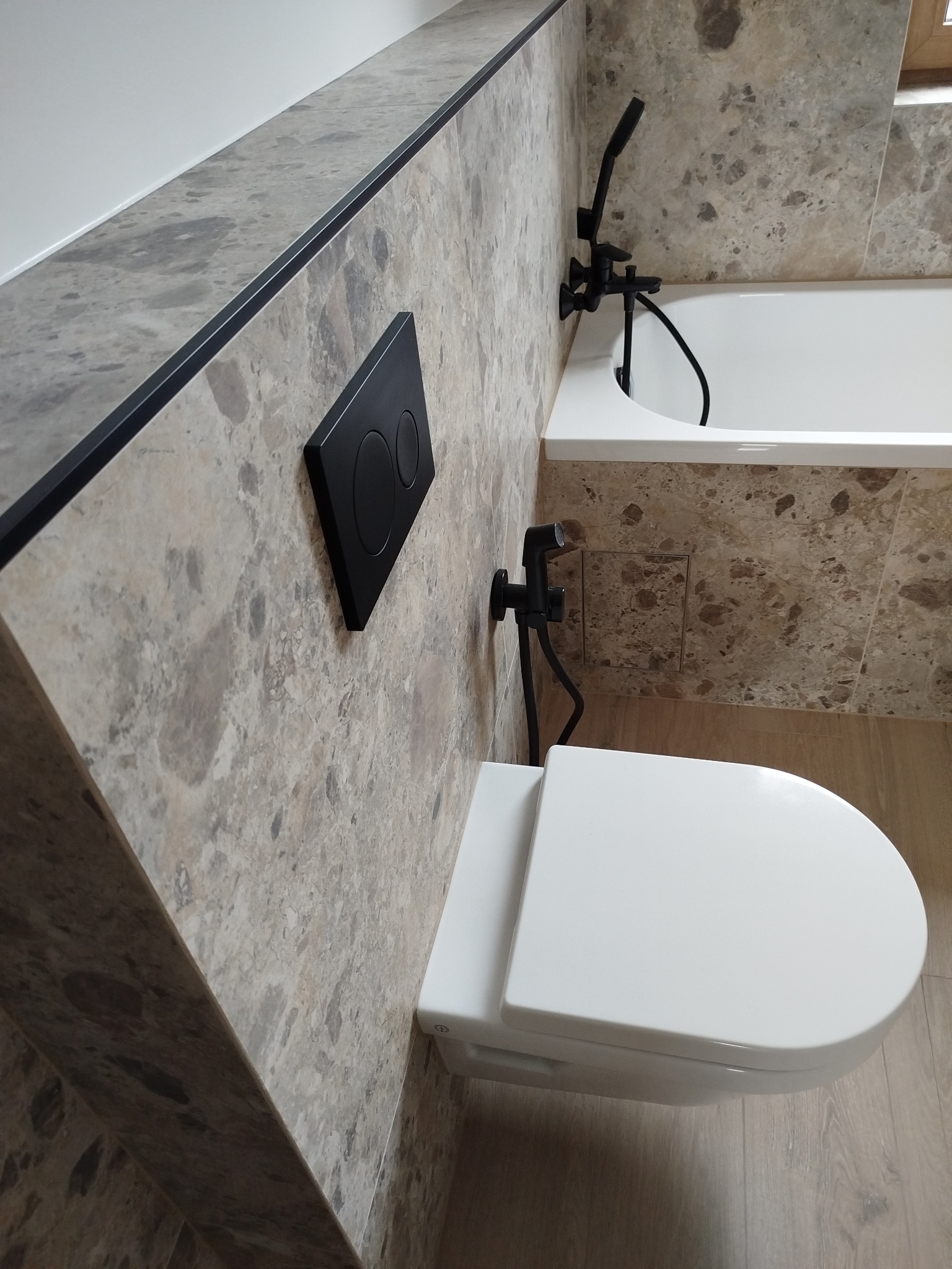 Wykończona łazienka z beżowymi płytkami imitującymi marmur, wisząca toaleta z czarnym przyciskiem spłukującym i czarna bateria bidetowa obok wanny.
