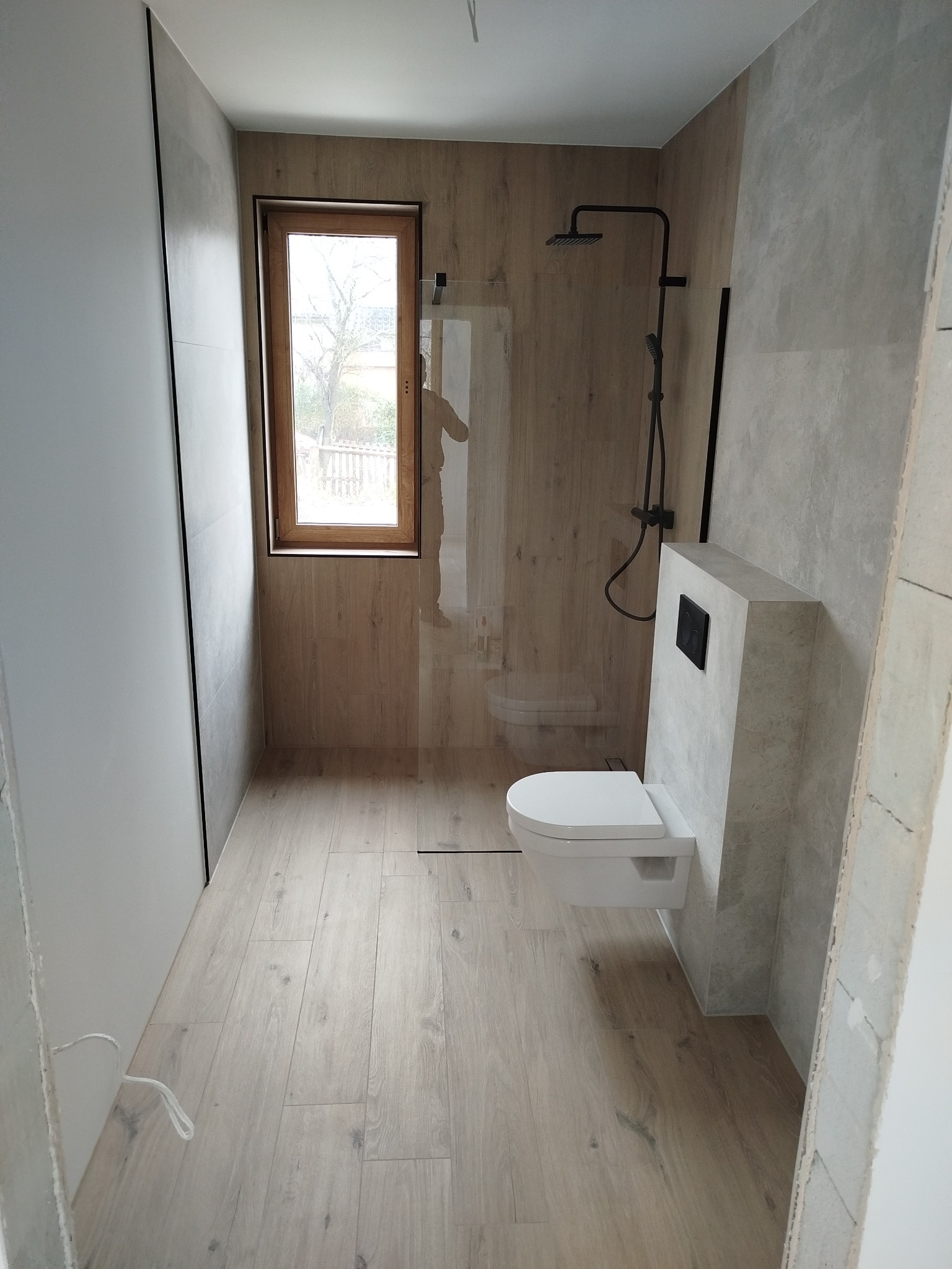 Nowoczesna łazienka z drewnopodobnymi płytkami, kabiną prysznicową typu walk-in i podwieszaną toaletą. Okno z drewnianą ramą w tle. Minimalistyczny design.