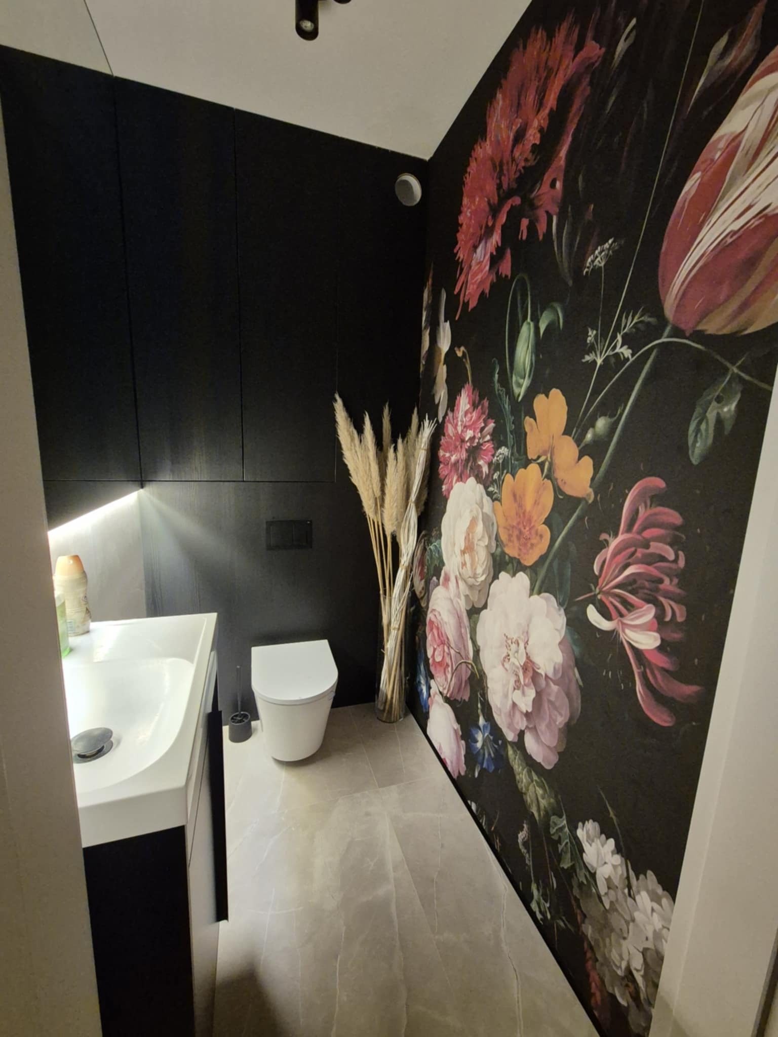Wykończona łazienka w Brzezinach: czarne ściany, umywalka z szafką, toaleta i fototapeta z motywem kwiatowym. Nowoczesny design i eleganckie wykończenie.