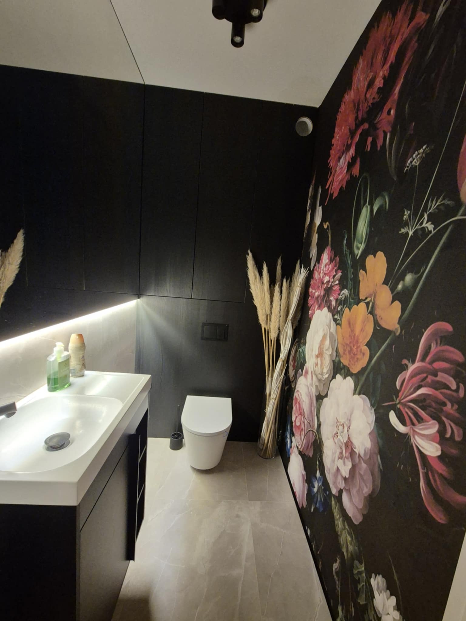 Nowoczesna łazienka z czarnymi szafkami, umywalką i toaletą. Ściana zdobiona tapetą w kwiaty, a całość dopełniają pampasowe trawy w wazonie.