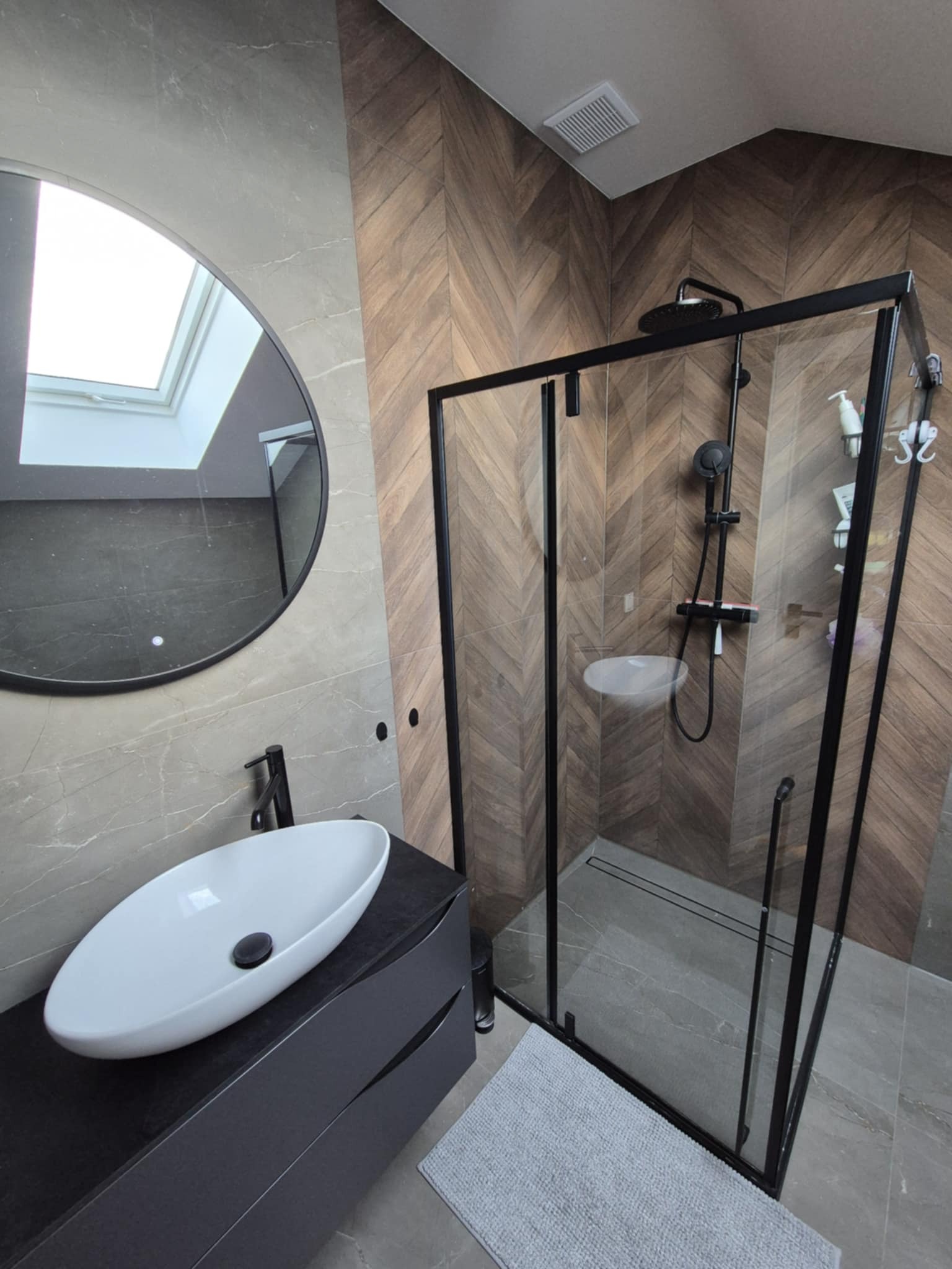 Nowoczesna łazienka z kabiną prysznicową typu walk-in z czarnymi profilami, umywalką nablatową i okrągłym lustrem. Ściany wykończone płytkami imitującymi drewno i marmur.
