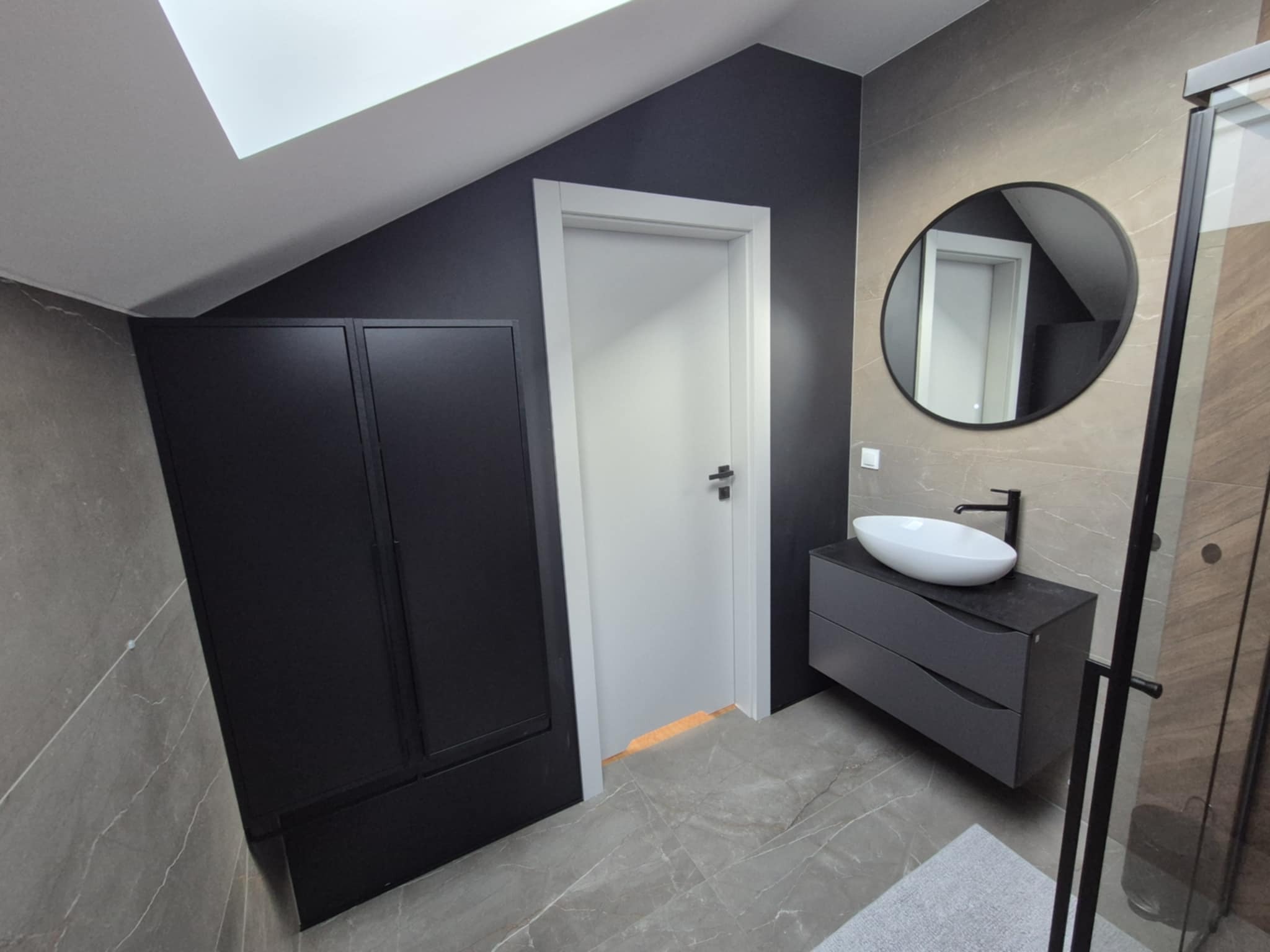 Nowoczesna łazienka na poddaszu z czarnymi akcentami: szafa, rama lustra i bateria. Umywalka owalna, biała. Płytki imitujące marmur. Kabina prysznicowa z czarną ramą.